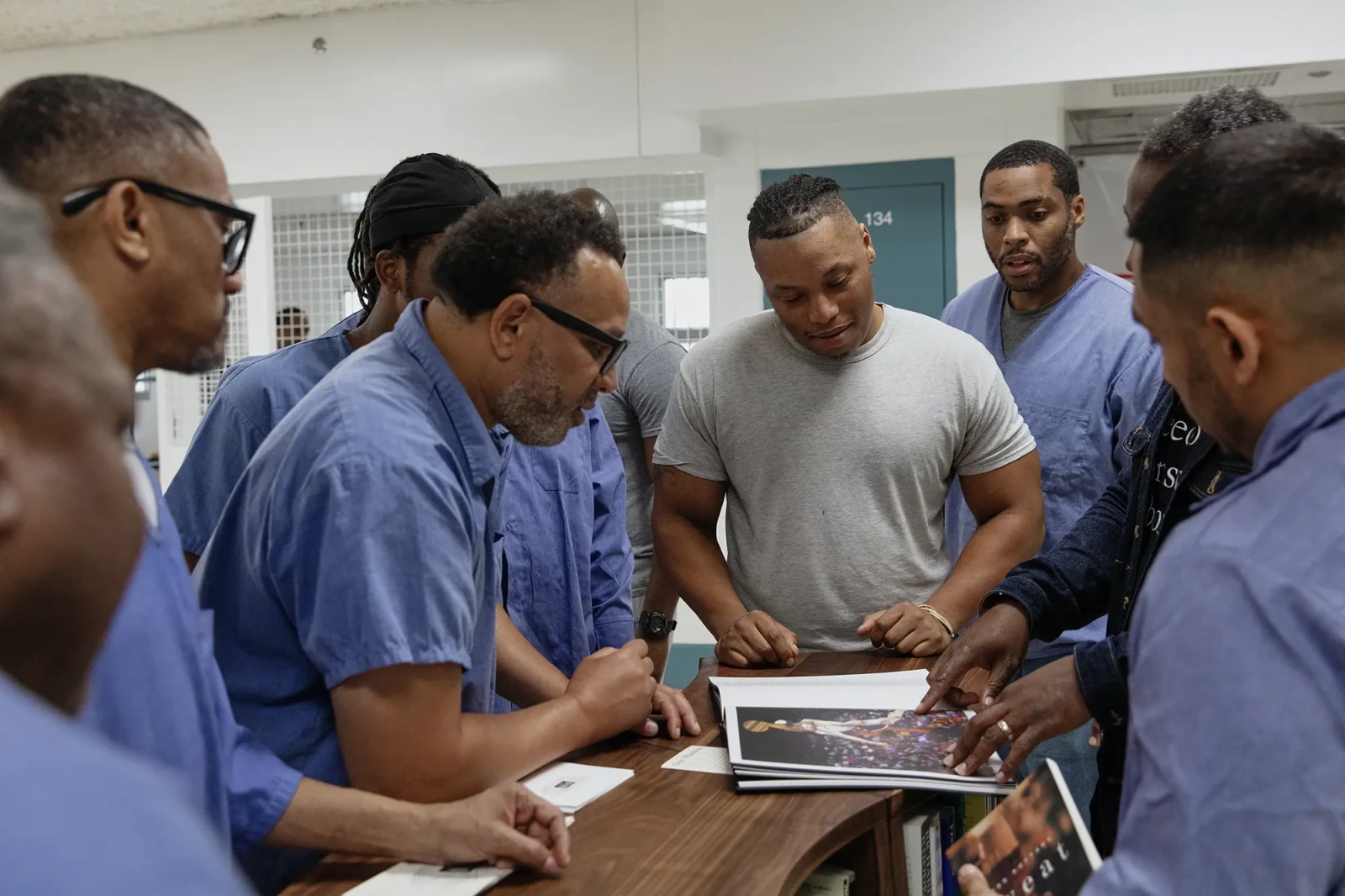 Freedom Library Patrons gather around the Freedom Library at Dorsey Run Correctional Facility in Maryland. Men clad in blue gather around a wooden Freedom Library bookcase discussing a copy of Reginald Dwayne Betts and Titus Kaphar’s Redaction book.