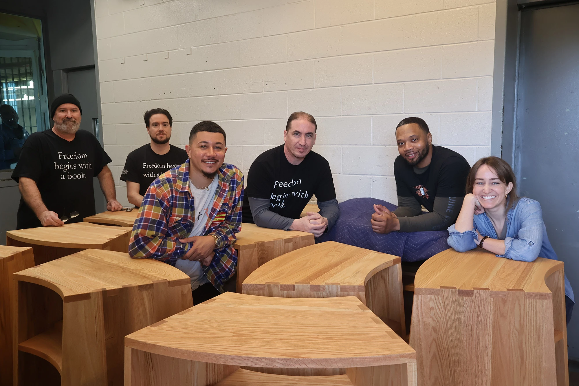 Freedom Reads Library Production Assistant Mike (second from right) and the Freedom Reads team with Freedom Libraries in Garden State Correctional Facility. 6 members of the Freedom Reads team pose for a photo around 9 wooden bookcases in a loading dock at Garden State Correctional Facility.
