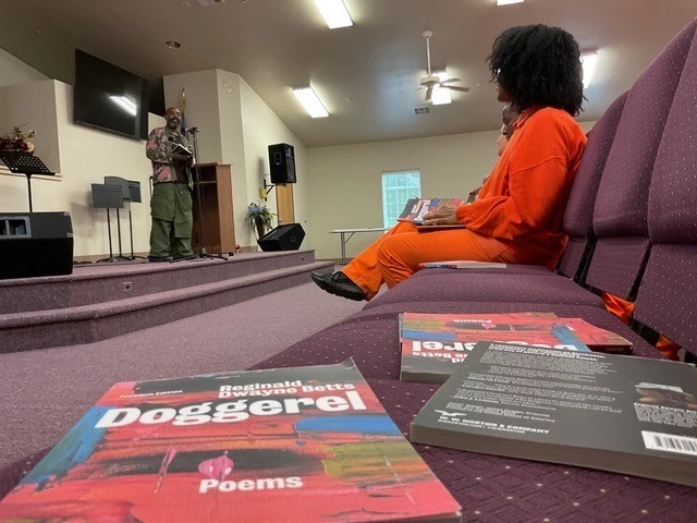 Freedom Reads Founder & CEO Reginald Dwayne Betts gave a reading from *Doggerel* at the Eddie Warrior Correctional Center for women. Watch (link: https://youtu.be/S8XYIQQIuTA?feature=shared text: a recap of Dwayne's reading) on YouTube. Doggerel books lay on auditorium chairs in a prison, women in the background in orange prison uniforms sitting watching Dwayne Betts perform on a small stage.