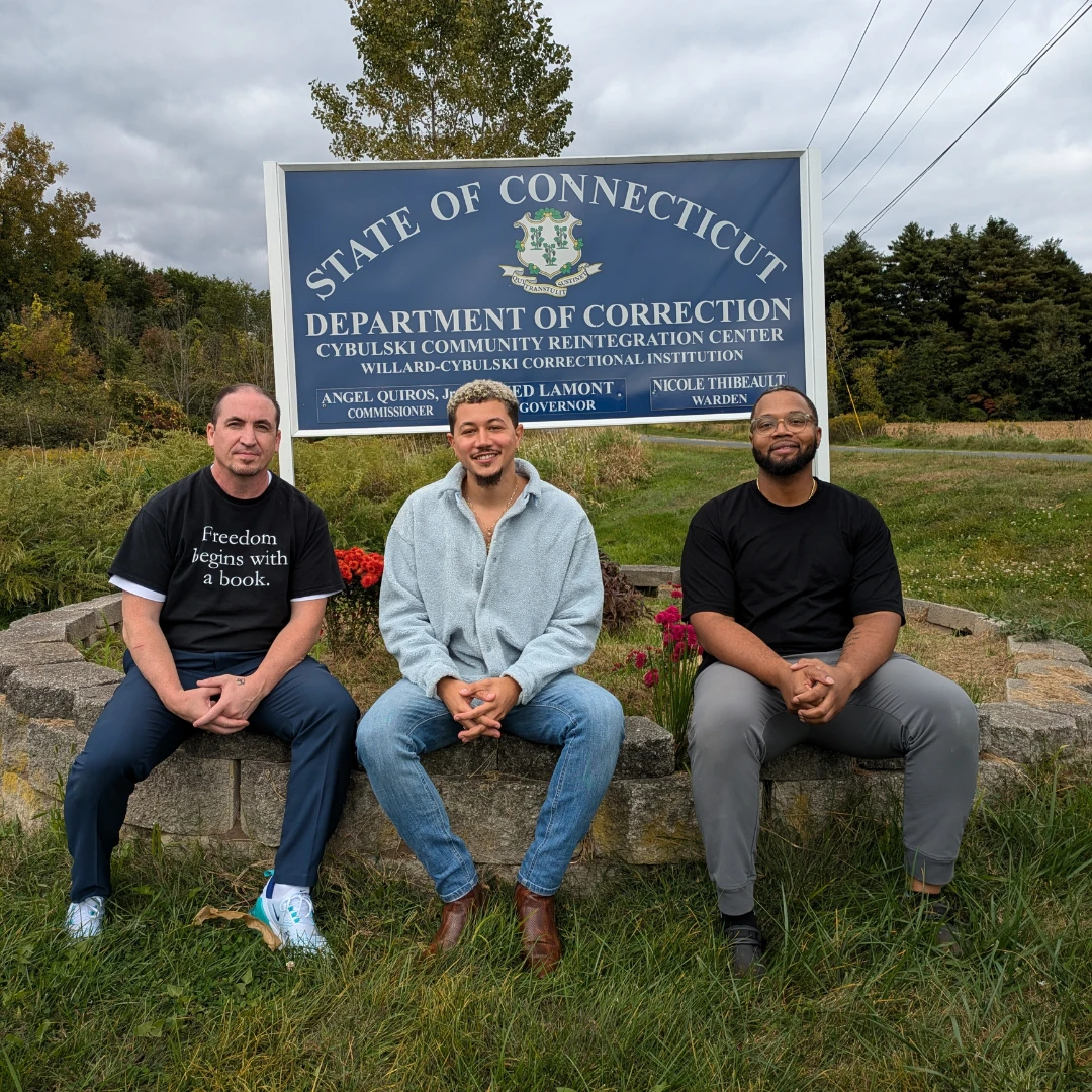 Freedom Reads Communications Manager Steven Parkhurst (left), Freedom Reads Library Coordinator David Perez Jr (center), and Freedom Reads Library Production Assistant Michael Byrd (right) sit outside of a sign for the Cybulski Community Reintegration Center.