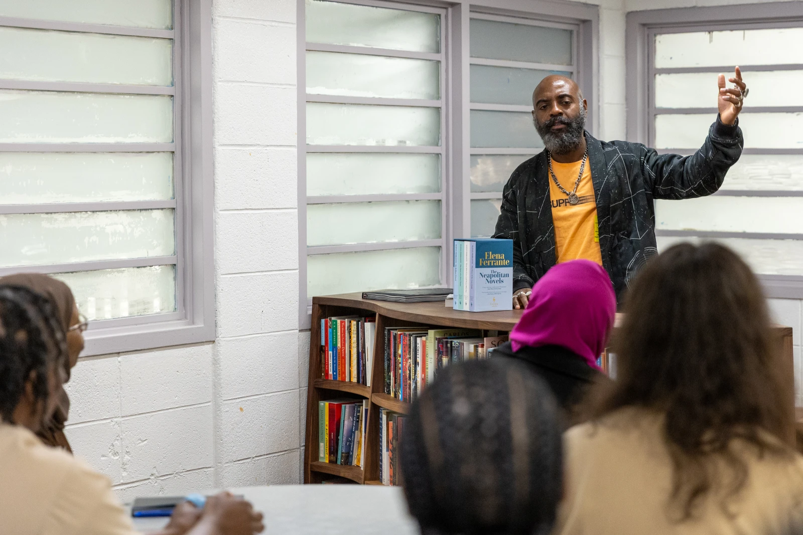 Freedom Reads Founder & CEO Reginald Dwayne Betts speaks at a Freedom Library opening at Edna Mahan Correctional Facility. Dwayne wearing a yellow shirt and black jacket speaking in front of newly opened freedom library wooden bookshelves at a women's prison